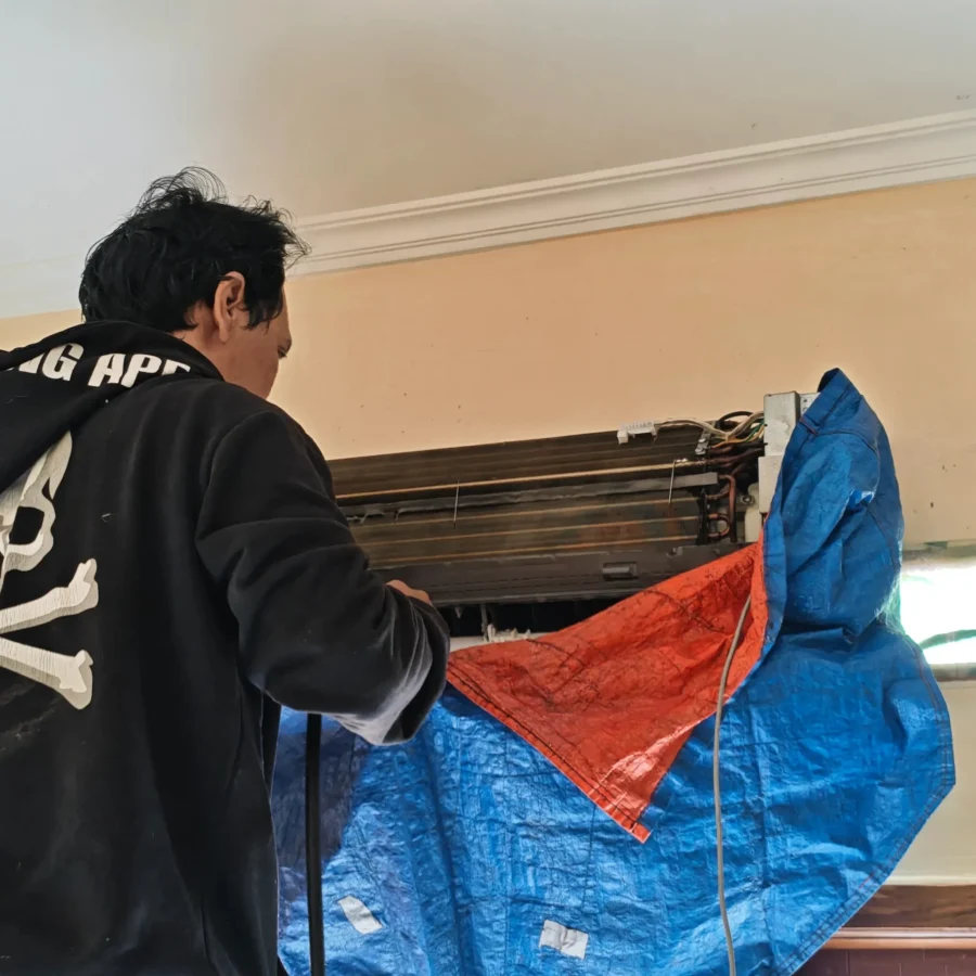 Seorang teknisi sedang melakukan proses pembersihan unit indoor AC yang kotor dan dipasangi terpal pelindung air